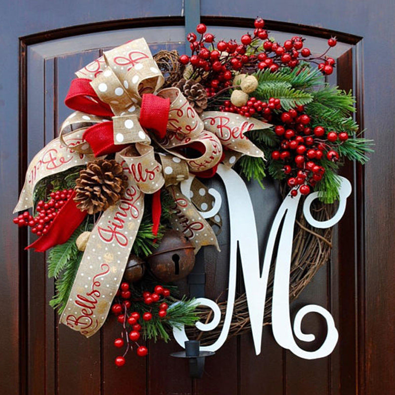 Festive wreath with pinecones, red berries, greenery, and a large red bow with "Jingle Bells" ribbon, featuring a white monogram "M" on a dark wooden door.