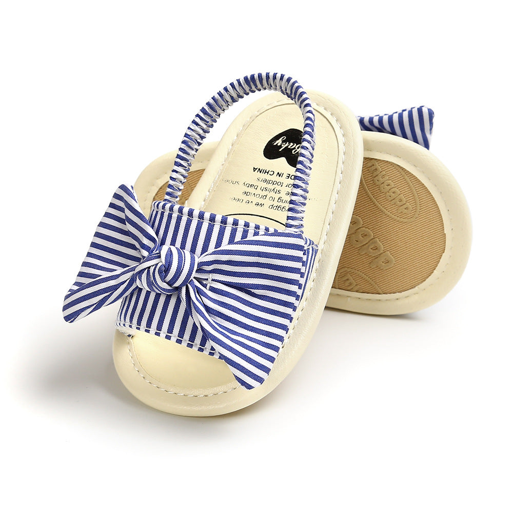 Striped baby shoes with a blue and white pattern, featuring an elastic strap and matching bow. Creamy toe cap and non-slip rubber sole visible.