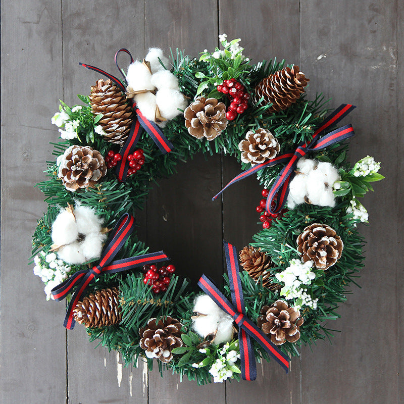 Festive wreath with pinecones, red berries, cotton sprigs, white flowers, and red-blue ribbons on green foliage, set against a rustic wooden background.