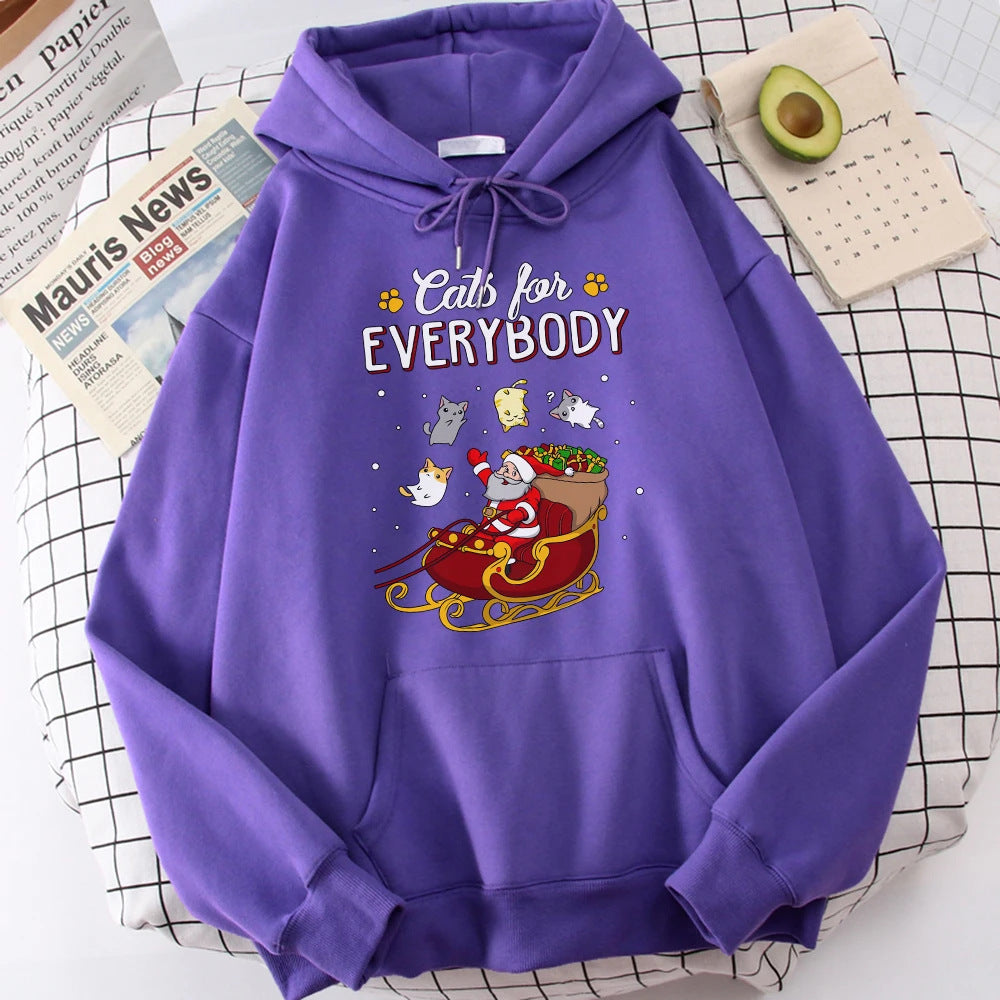 Purple hoodie with "Cats for Everybody" slogan featuring a playful Santa in a sleigh surrounded by floating cats. Background shows a newspaper, grid paper, and half an avocado.