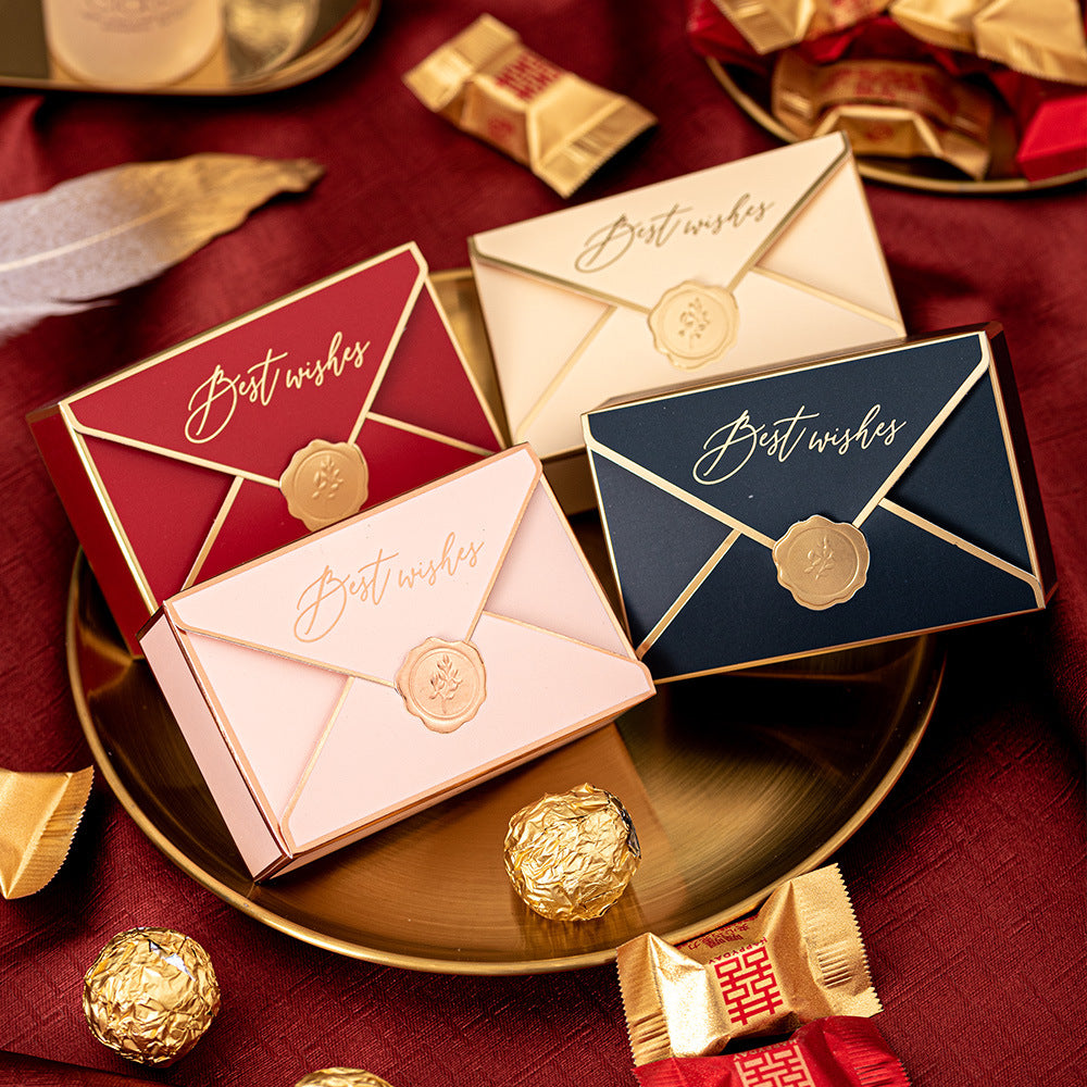 Gift boxes resembling envelopes with "Best wishes" text, in red, pink, cream, and navy, with gold seals on a tray, surrounded by chocolates and wrapped treats on a red fabric.