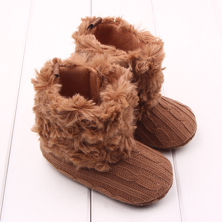 Brown baby booties with fluffy faux fur cuffs and cable-knit pattern on a white wooden floor. Cozy and stylish, perfect for keeping tiny feet warm.