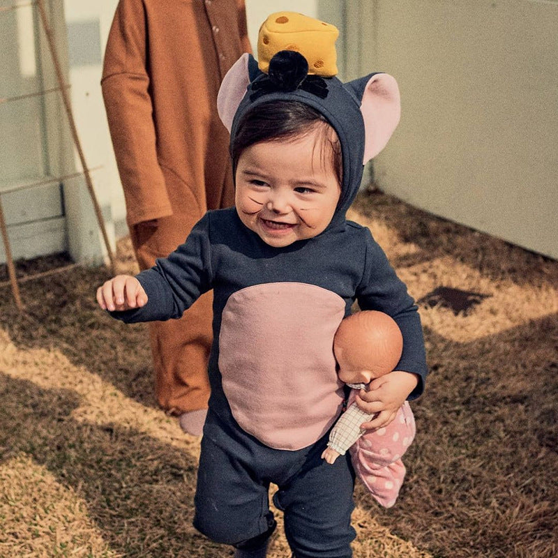 A child in a mouse costume with a cheese hat smiles while holding a doll. The child is outdoors on grass, and another person in a brown outfit is partially visible in the background.