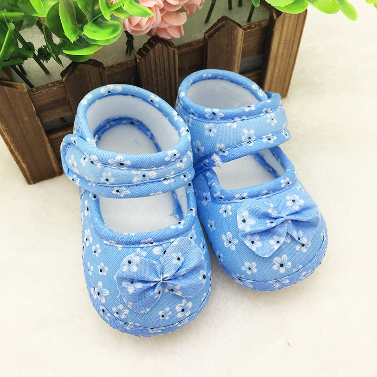 Blue baby shoes with white floral patterns and bows, set against a wooden fence with green leaves and pink flowers in the background.