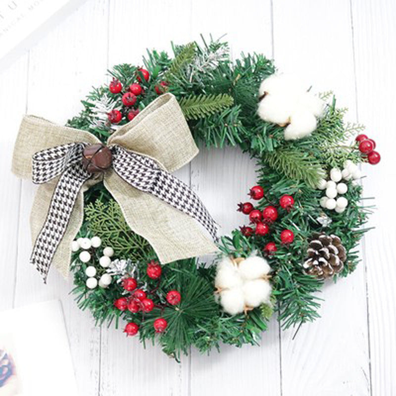 A festive wreath with green pine branches, red and white berries, a pinecone, cotton accents, and a burlap bow with a black and white ribbon, on a white wooden background.
