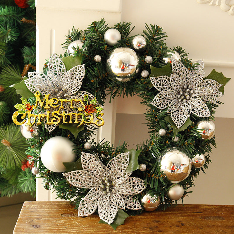 Christmas wreath with silver ornaments, glittery silver poinsettias, and green foliage, featuring a "Merry Christmas" sign. Displayed on a wooden table near a Christmas tree.
