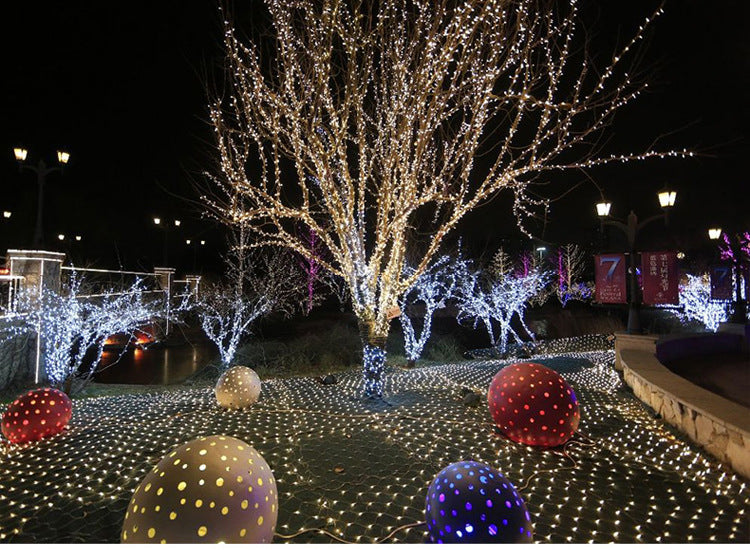 A tree adorned with sparkling white lights stands among other illuminated trees, surrounded by large, colorful polka-dotted spheres on a ground covered in string lights.
