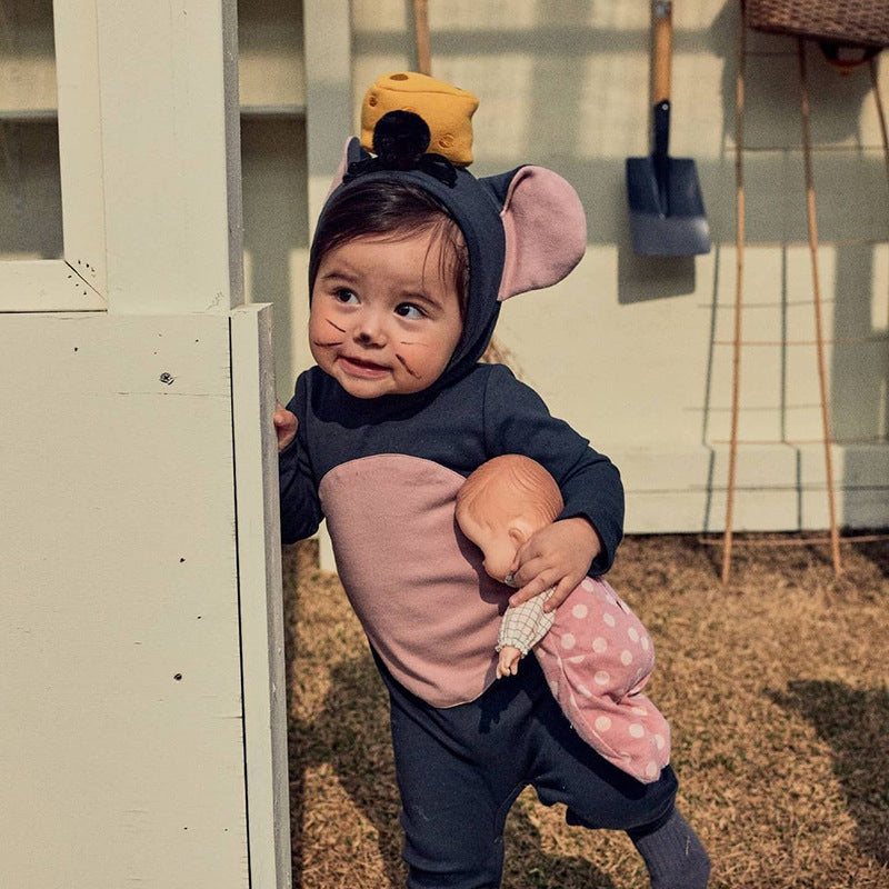 A toddler in a cute mouse costume with pink ears and a cheese accessory on the hood holds a baby doll while peeking from behind a white structure outdoors.