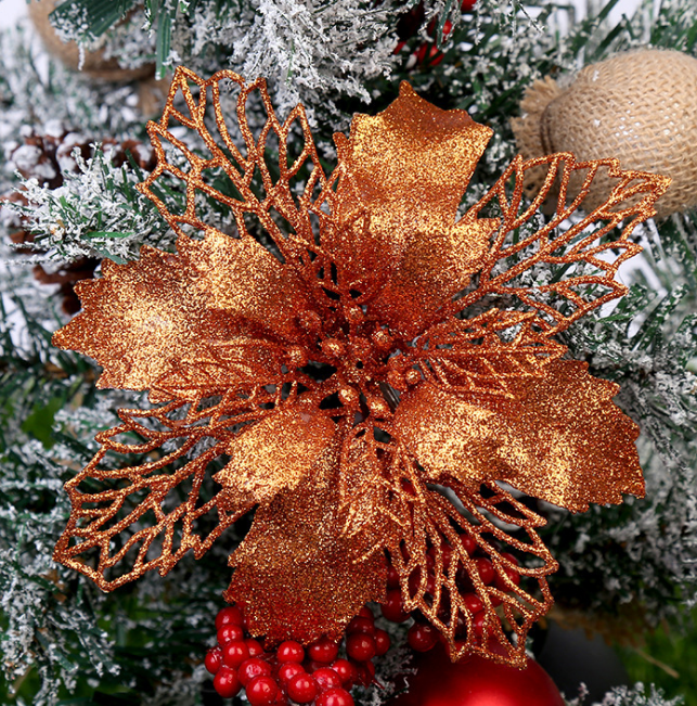 Glittery orange poinsettia ornament on a snow-frosted Christmas tree with red berries and burlap accents, creating a festive holiday decoration.