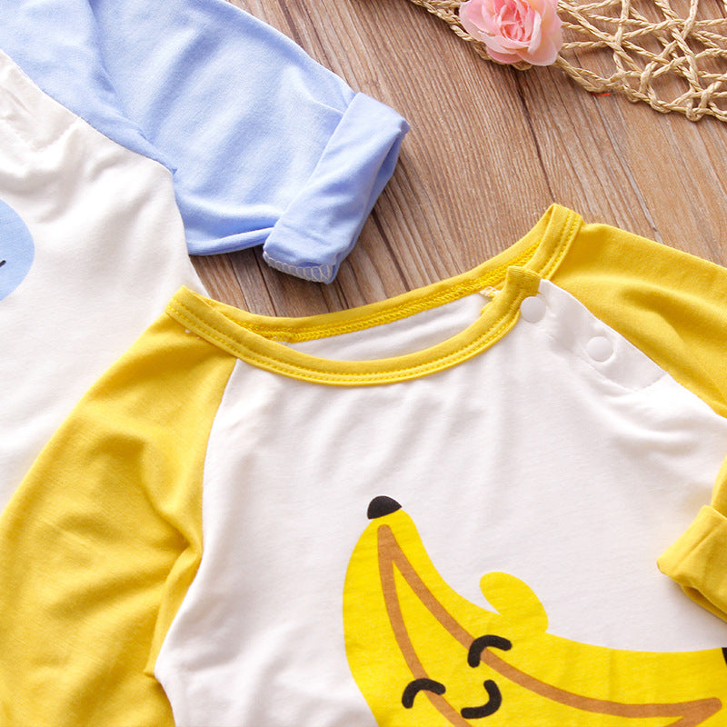Two children's shirts lie on a wooden surface; one has yellow sleeves with a smiley banana print, the other is partially visible with light blue sleeves. A pink rose and woven mat are nearby.