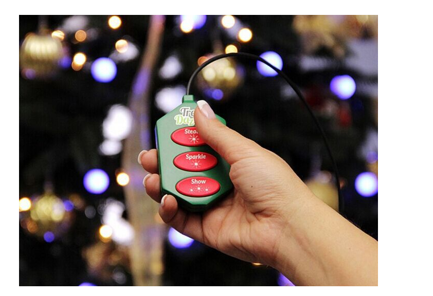 A hand holds a green and red remote with buttons labeled "Steady," "Sparkle," and "Show," in front of a lit Christmas tree with blurred lights in the background.