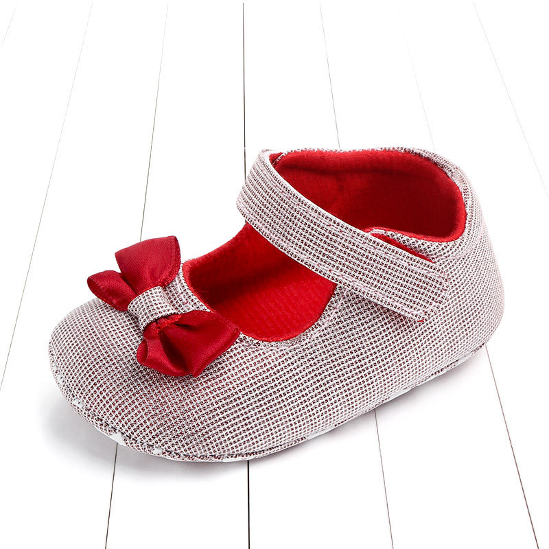 A baby shoe with a red interior and bow, featuring a silver textured pattern and a Velcro strap, is placed on a white wooden floor.