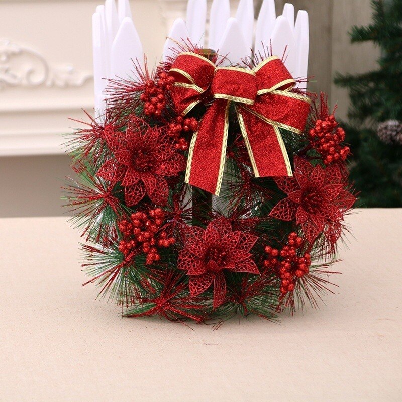 A festive wreath with red poinsettias, berries, and a large red bow, displayed indoors on a beige surface, with a hint of a Christmas tree in the background.