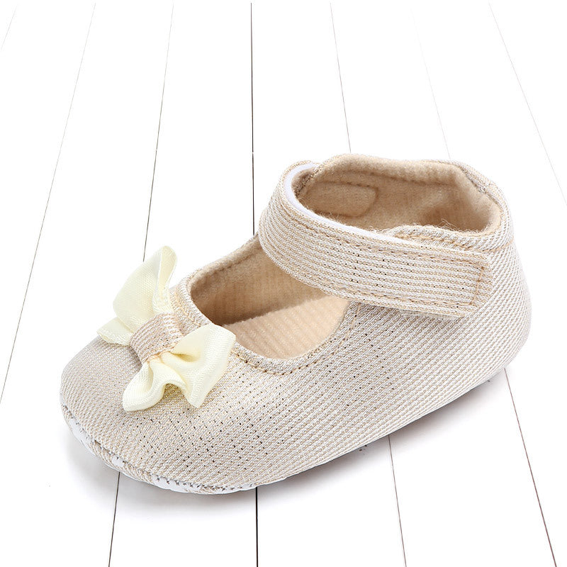 Beige baby shoe on a white wooden surface, featuring a soft fabric texture, an ankle strap closure, and adorned with a cream-colored bow on the front.