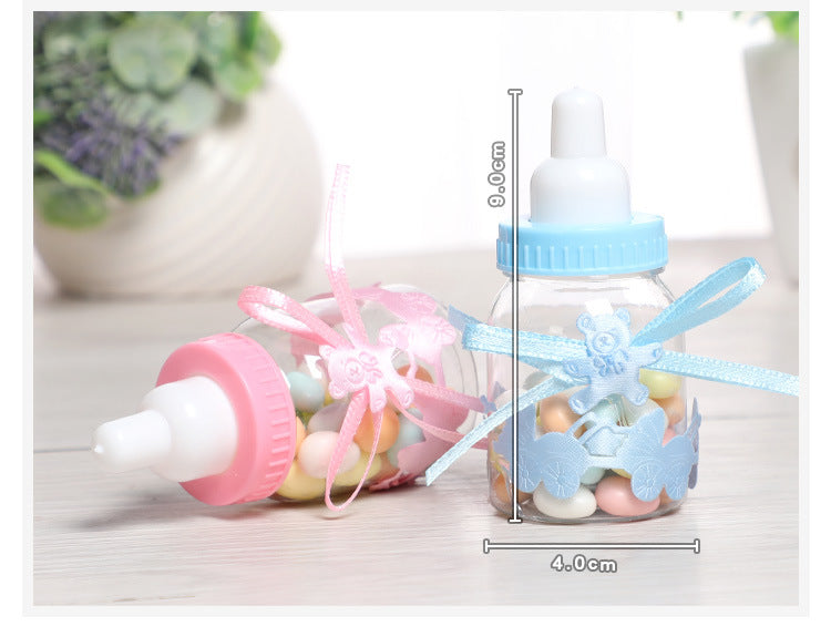 Two small plastic baby bottles filled with colorful candy. One bottle has a pink cap and ribbon, the other blue. Dimensions shown: 9 cm tall, 4 cm wide. Background has a blurred plant.