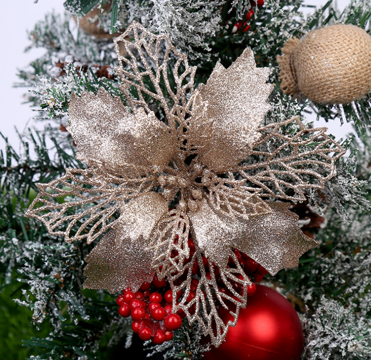 Glittery golden poinsettia on a snowy Christmas tree branch, adorned with red berries, a burlap ornament, and a shiny red bauble, creating a festive holiday scene.