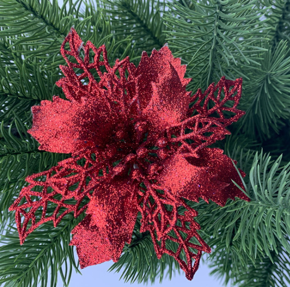 A vibrant, glittery red poinsettia ornament with intricate leaf patterns is nestled among lush green pine branches, creating a festive holiday decoration.