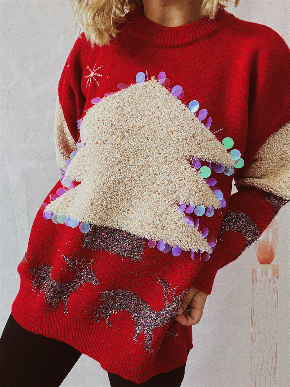 A person wearing a festive red sweater with a large 3D Christmas tree design, adorned with colorful sequins and reindeer patterns, stands in front of a neutral background.