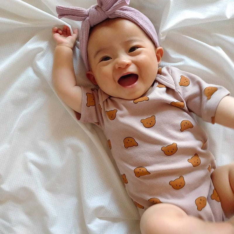 Smiling baby wearing a pink headband and a light pink onesie with brown teddy bear prints, lying on a white blanket.