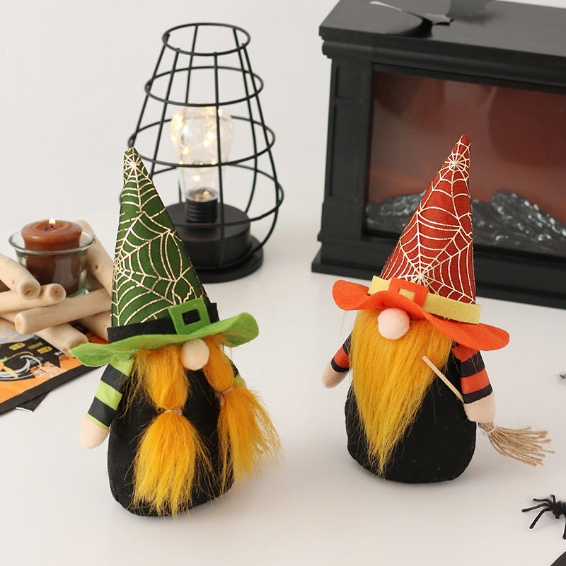 Two plush gnomes with yellow beards and conical hats, one green and one orange, featuring spiderweb patterns. They stand on a table with Halloween decor, including a candle, a black wire lamp, and a decorative fireplace. A tiny broom and spider accent the scene.