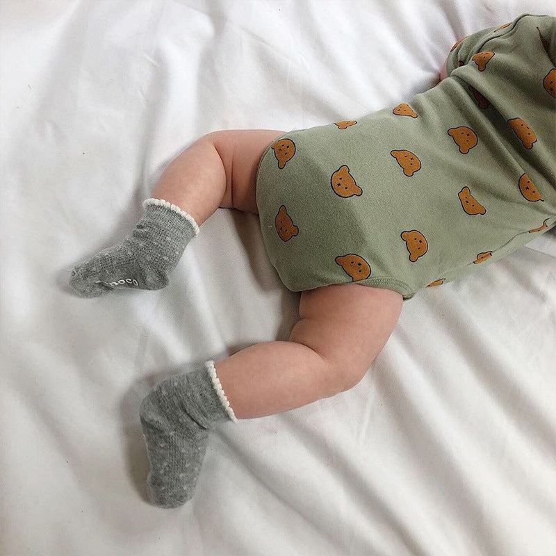 A baby lying on a white bed, wearing a green onesie with bear face patterns and grey socks with white trim.