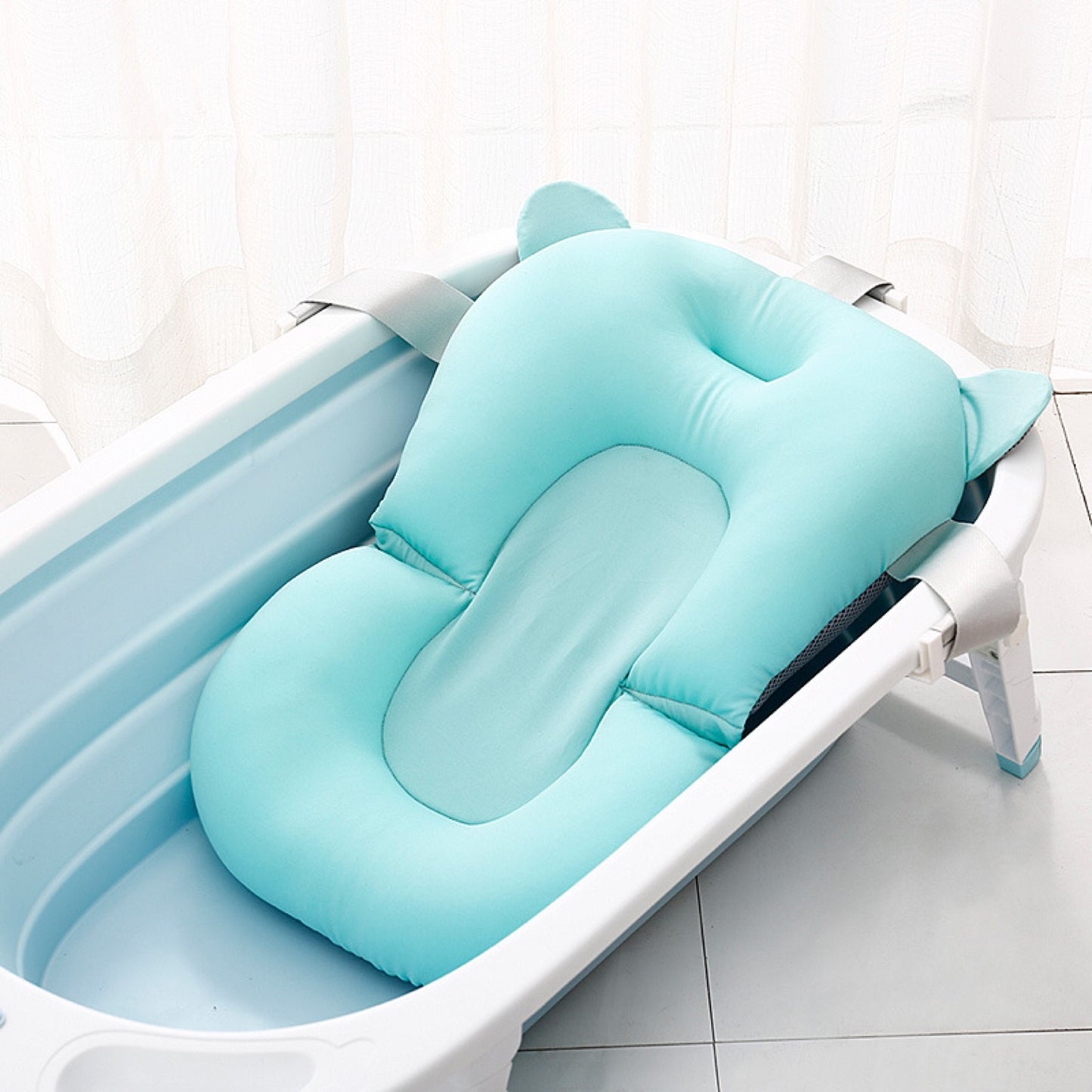 A light blue baby bathtub with a soft, cushioned insert featuring cute ear-like designs. The tub is placed on a tiled floor with white curtains in the background.