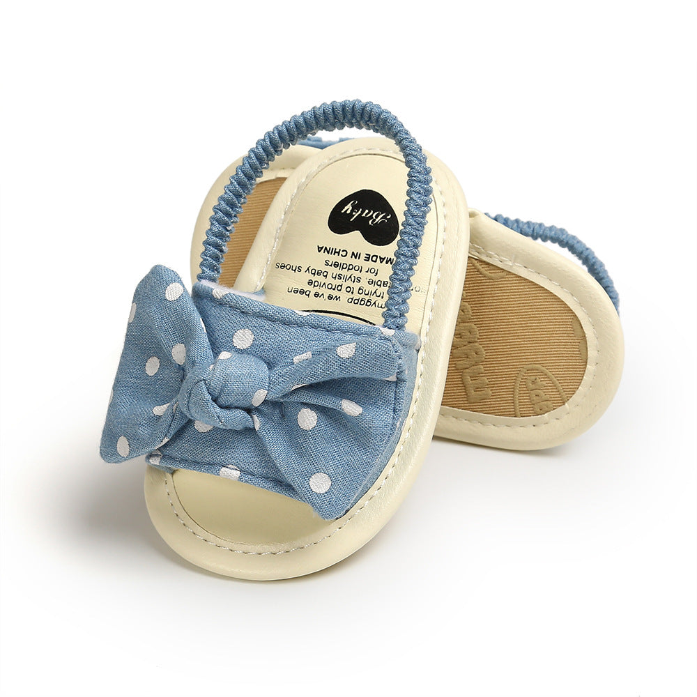 Baby sandals with blue polka dot bows and elastic straps. Cream soles with text inside one sandal. Solid construction, designed for comfort and style.