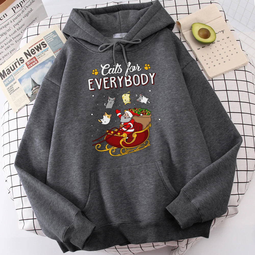 Gray hoodie with "Cats for Everybody" text features Santa in a sleigh surrounded by playful cats. Nearby, a newspaper and half an avocado on a checkered surface.