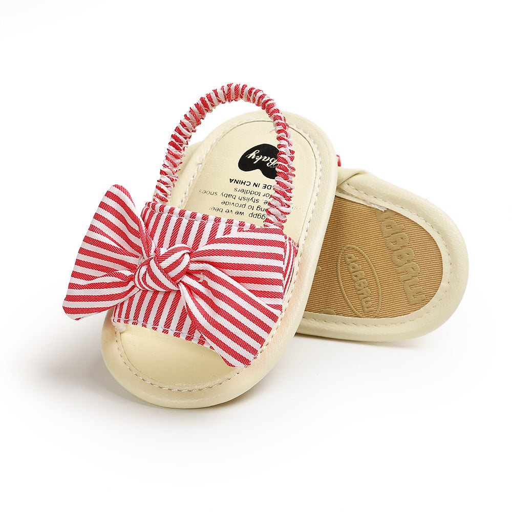 Baby sandals with red and white striped fabric bows and elastic straps. Cream-colored base with a textured sole, designed for infant comfort and style.