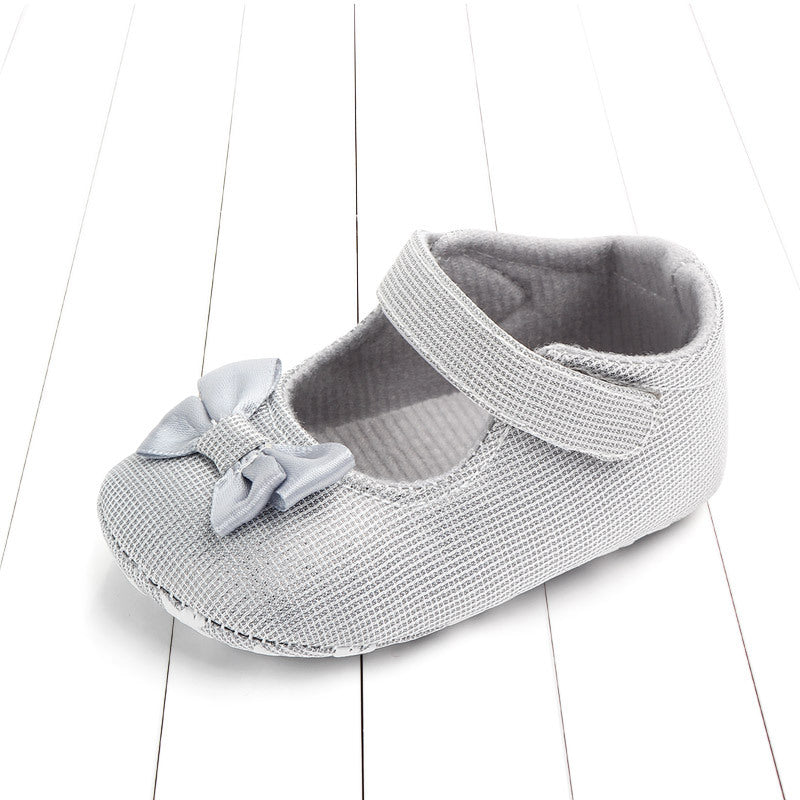 Silver baby shoe with a Velcro strap and a decorative bow on the toe, placed on a white wooden floor. The shoe has a textured, sparkly surface.