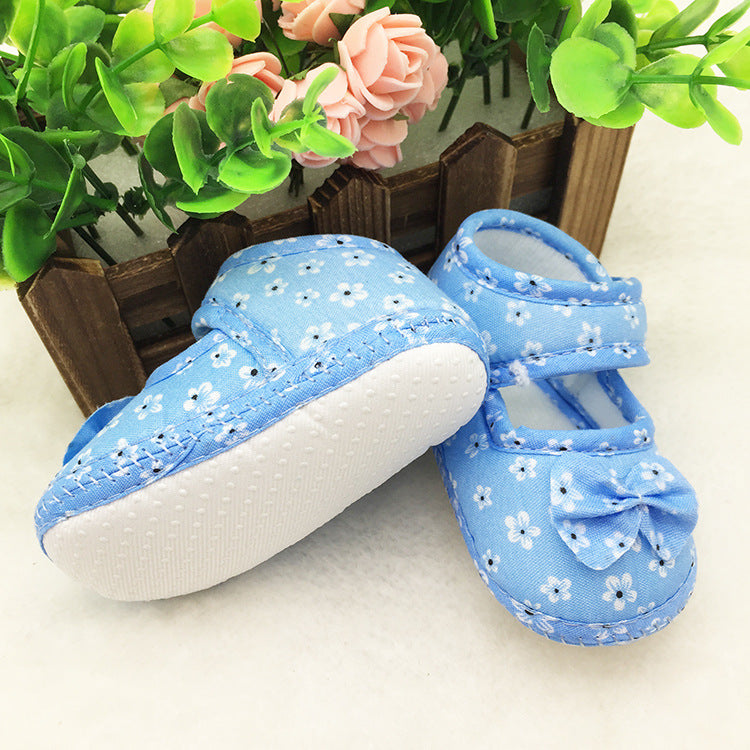 Blue baby shoes with white floral patterns and bows, placed near pink and green artificial flowers in a wooden planter box on a soft white surface.
