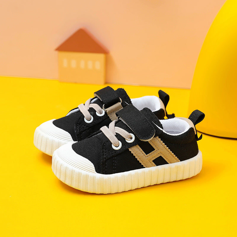 Black and white toddler sneakers with beige accents and Velcro straps, set against a bright yellow and peach backdrop with a small house decoration in the background.