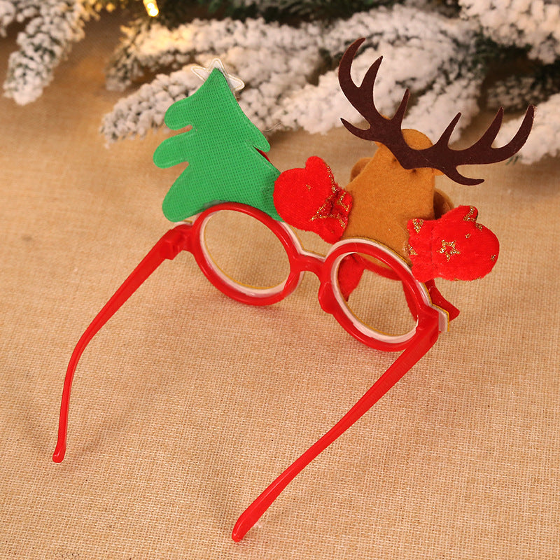 Festive eyeglasses with red frames, decorated with a green Christmas tree on one side and a brown reindeer with antlers on the other, set against a snowy pine backdrop.