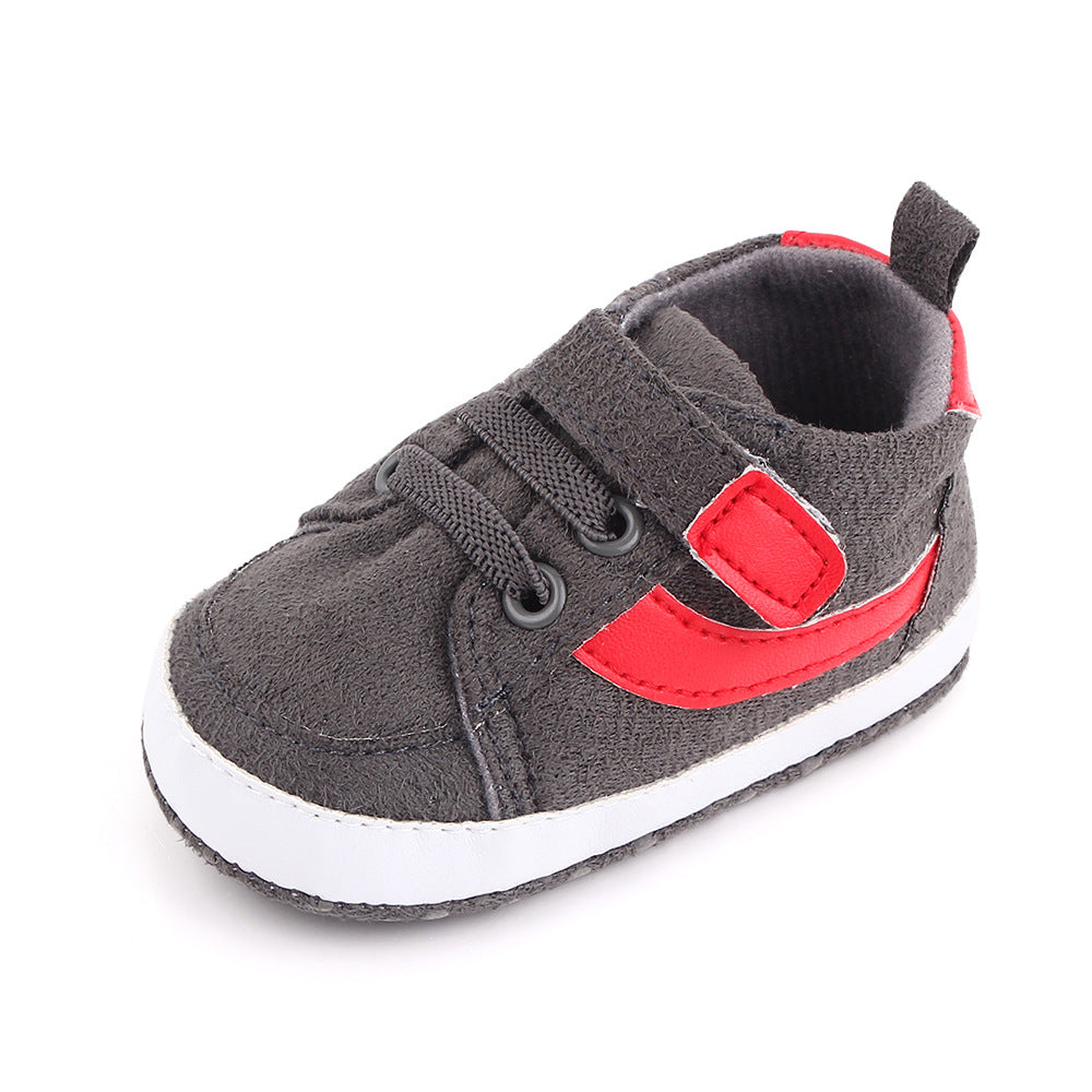 Gray baby sneaker with black laces, featuring a red strap and accent on the side. The shoe has a white sole and a pull tab at the heel for easy wearing.