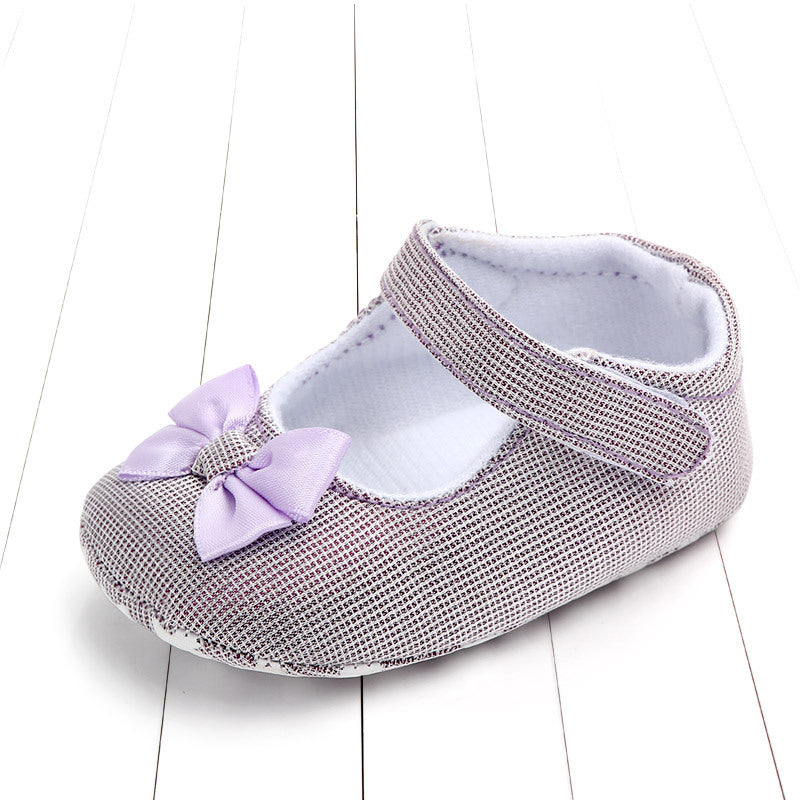 Baby shoe with a purple checkered design, featuring a soft lilac bow on the front and an adjustable strap. The shoe is displayed on a white wooden surface.