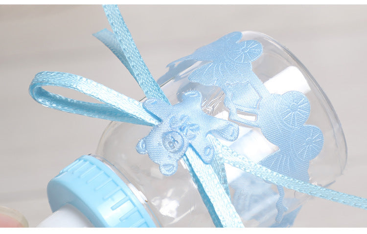 A close-up of a transparent baby bottle adorned with a blue ribbon and bear decoration. The bottle cap is light blue, and a lace-like pattern decorates the outer surface.