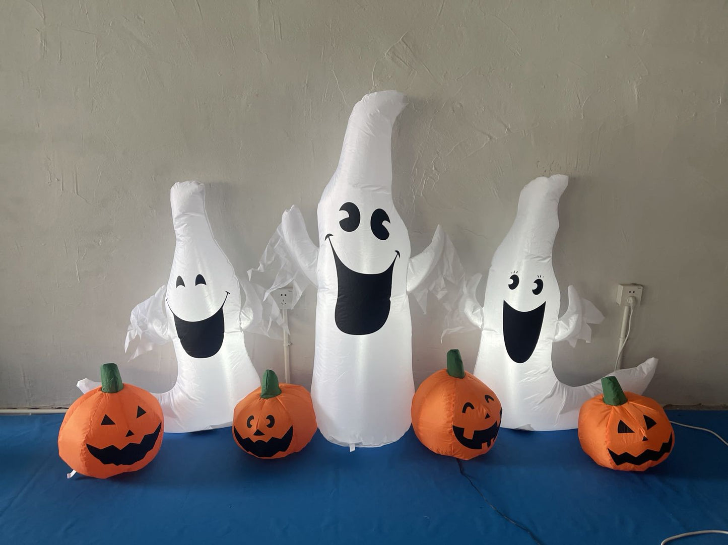 Three inflatable ghosts with cheerful faces are surrounded by five inflatable pumpkins with carved expressions, set against a plain wall and a blue floor.