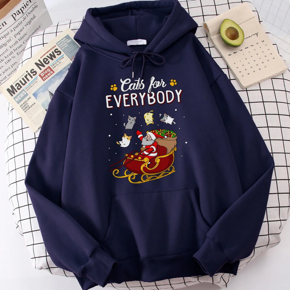 Navy hoodie on a grid-pattern surface, featuring Santa in a sleigh with cats, surrounded by the text "Cats for Everybody." Nearby, an avocado, a newspaper, and a calendar are visible.
