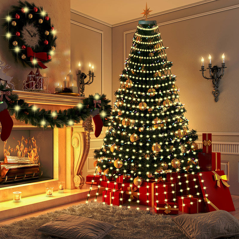 A cozy living room with a decorated Christmas tree adorned with lights and gold ornaments. A fireplace is lit, with stockings hanging, and a wreath above. Gifts are placed under the tree.