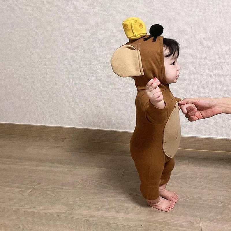 A toddler in a brown mouse costume with a yellow cheese hat stands on a wooden floor, held by an adult's hand for support. The child looks to the right.
