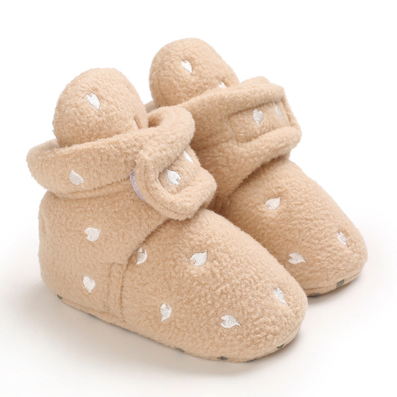 Cozy baby booties in beige, featuring a soft, fluffy texture with small white heart patterns. Velcro straps ensure a snug fit, perfect for keeping tiny feet warm.
