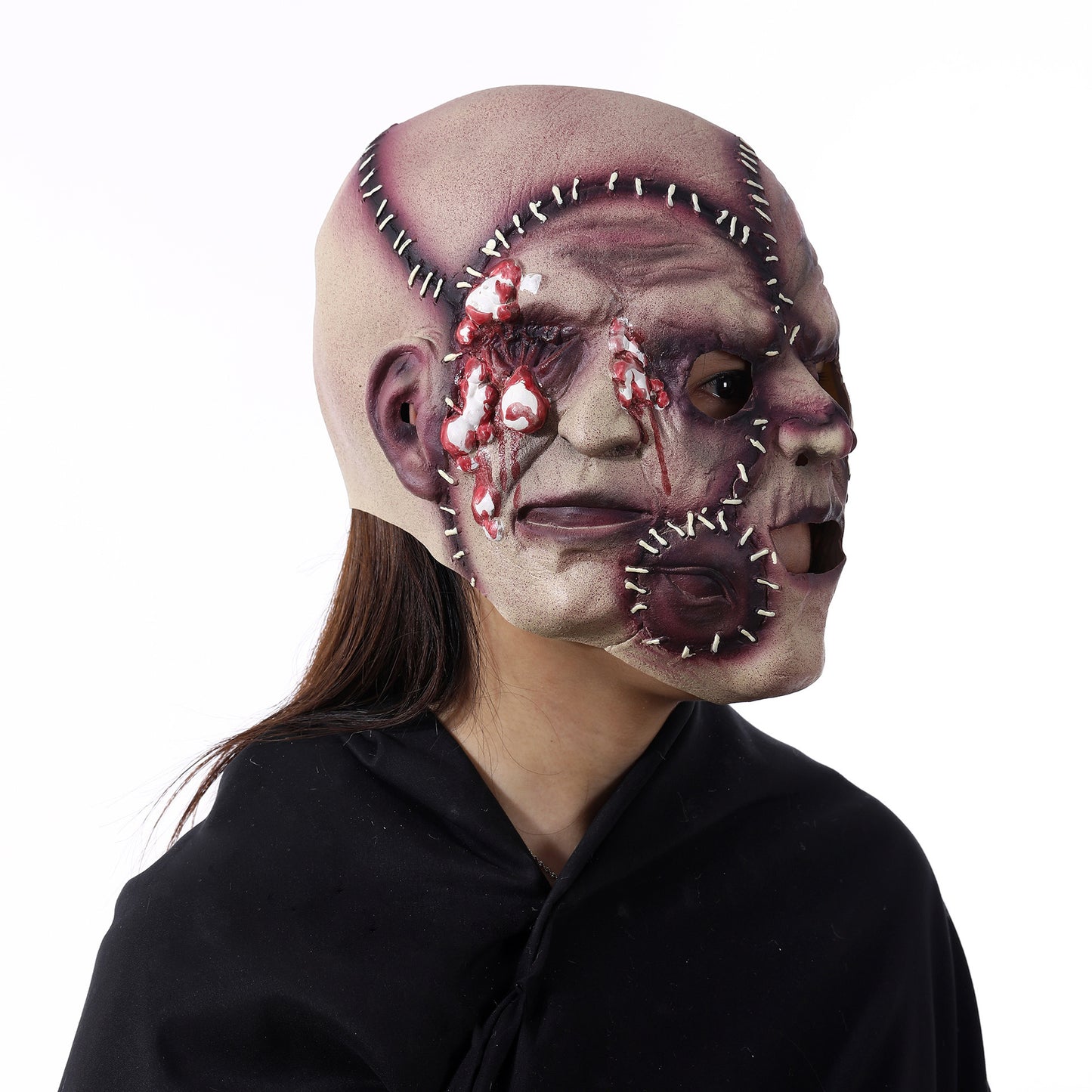 Person wearing a horror mask with sewn seams and gruesome details, including multiple faces and dripping blood. Dark hair and black cloak enhance the unsettling effect.