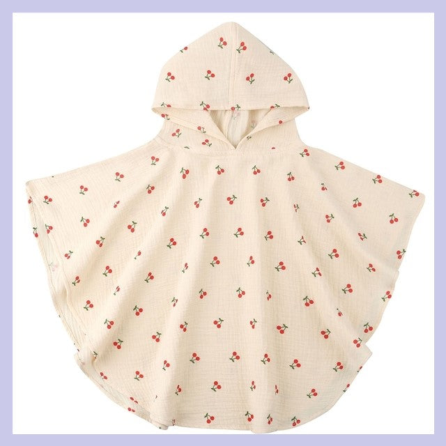 Cream-colored hooded cape for children with a cherry pattern, featuring small red and green motifs on a soft fabric. The cape is set against a light purple background.
