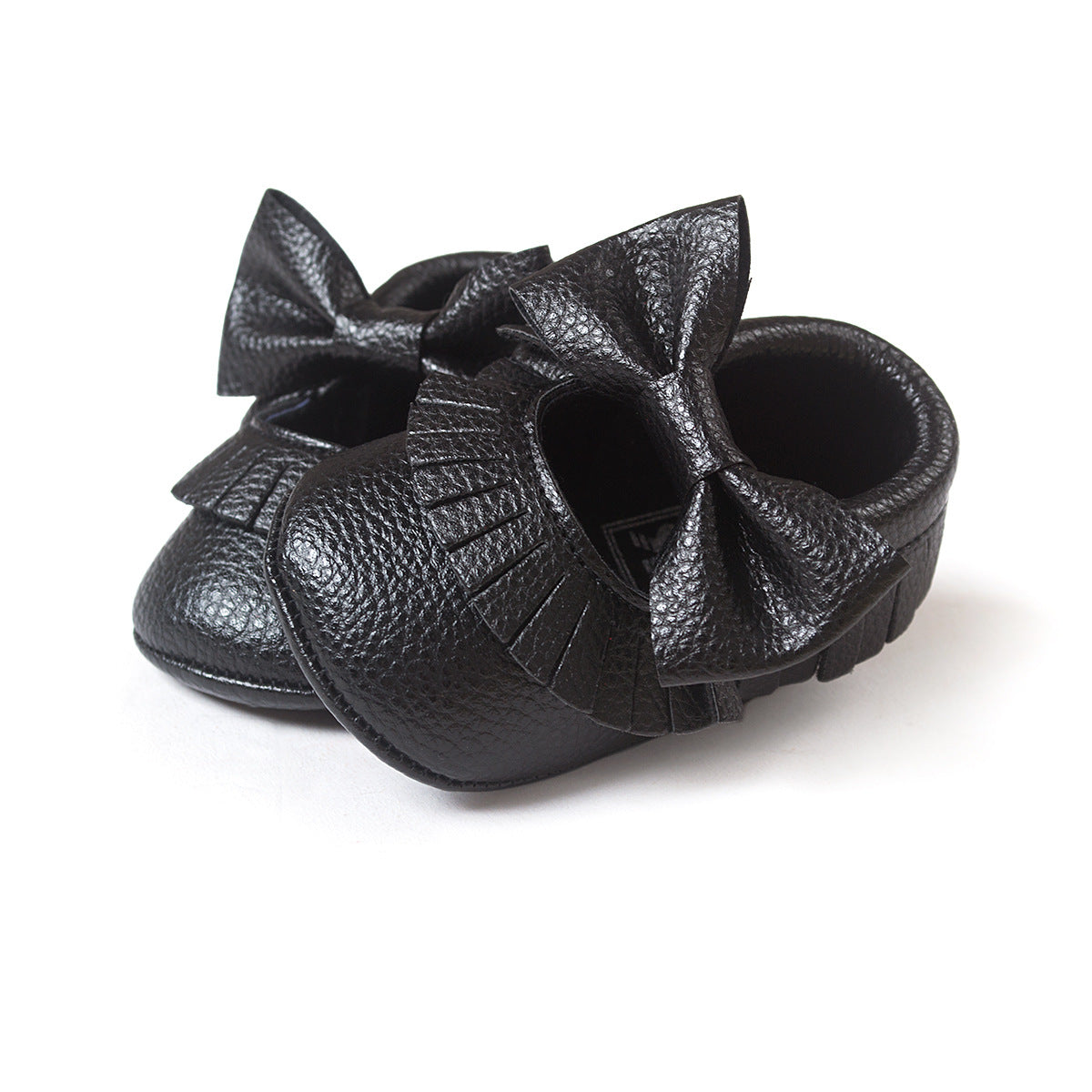 Black baby shoes with textured leather and large bows on top. The shoes have a decorative fringe around the opening, set against a white background.