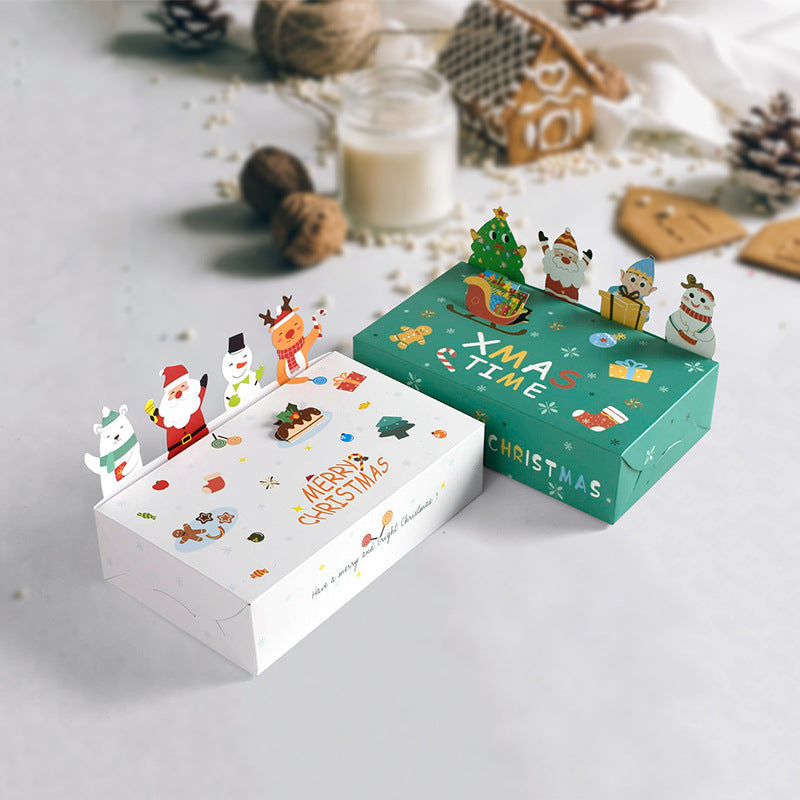 Two festive gift boxes on a table, one white and one green, decorated with Christmas-themed graphics and cutout figures like Santa and a snowman. Background includes holiday items.