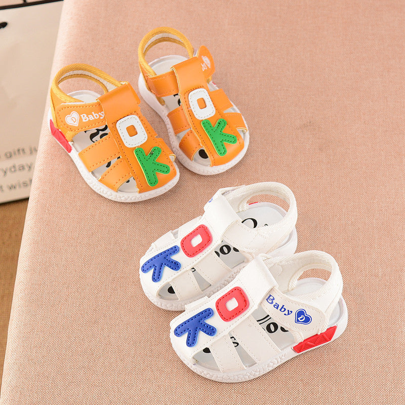 Two pairs of colorful baby sandals on a soft pink surface. One pair is orange with green and white letters, the other is white with blue, red, and pink letters.