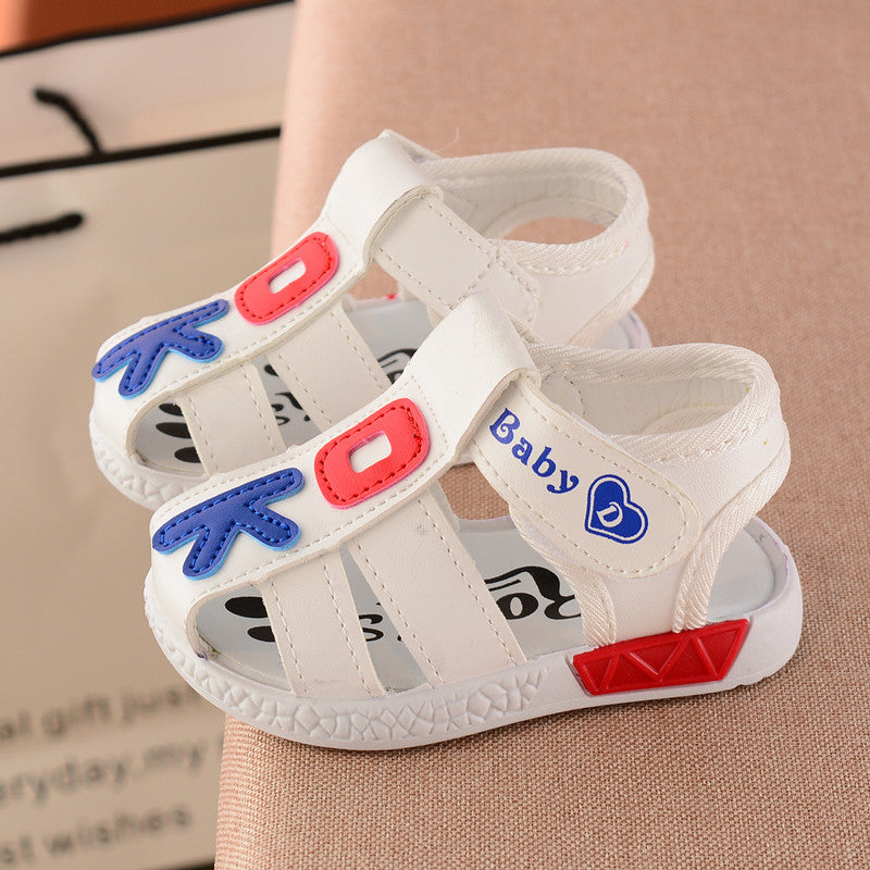 White baby sandals with red and blue accents, featuring "Baby" text and heart symbol. The sandals have a cushioned sole and open-toe design on a beige background.