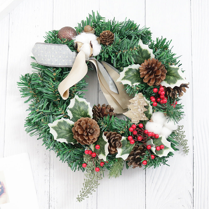 Festive wreath with pinecones, red berries, cotton, and green foliage, accented by a beige and silver ribbon on a white wooden background.