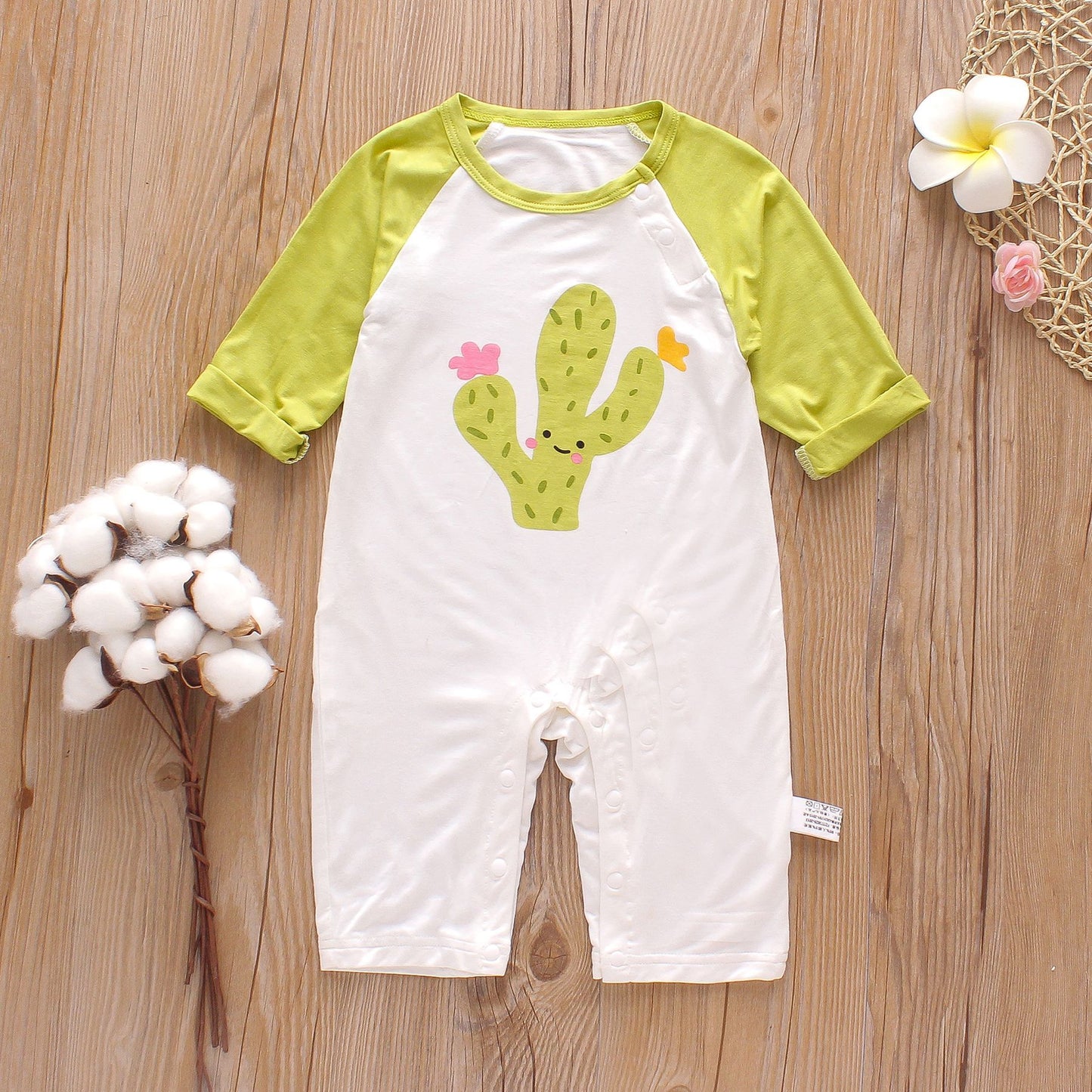 Baby romper with green sleeves and a smiling cactus print on white fabric, placed on a wooden surface. Nearby are cotton flowers and decorative beige and white elements.