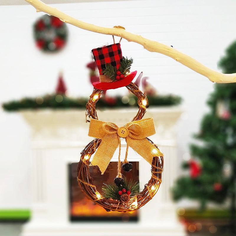 Rustic Christmas wreath shaped like a snowman with a buffalo plaid top hat, burlap bow, and pine embellishments, illuminated with fairy lights, hangs in front of a festive fireplace.